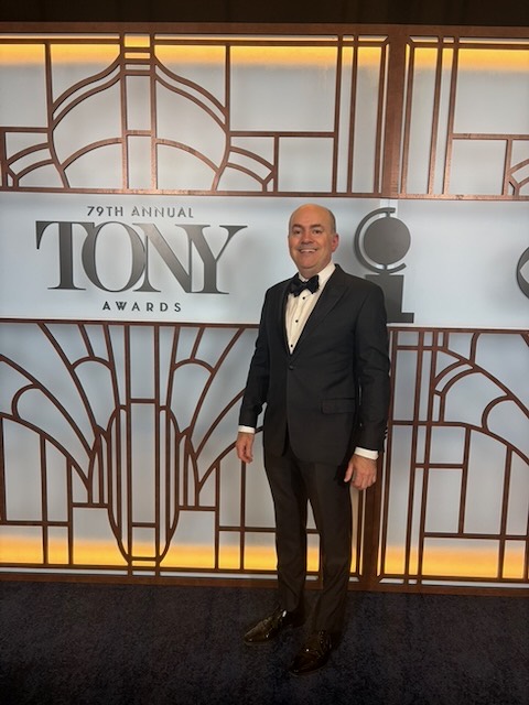 Brian Guy in a tuxedo on the blue carpet at the 2026 Tony Awards, in front of the 79th Annual Tony Awards art-deco backdrop